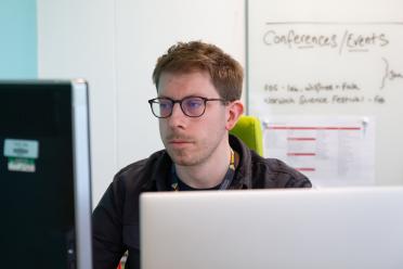 Photo of Greg, who has dark blonde hair and wears brown glasses, working at his desk with two screens.