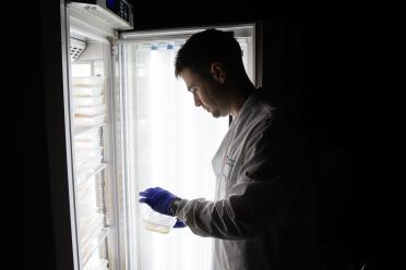 PhD researcher Jonathan photographed in low light in front of an open brightly lit refrigerator full of seed samples