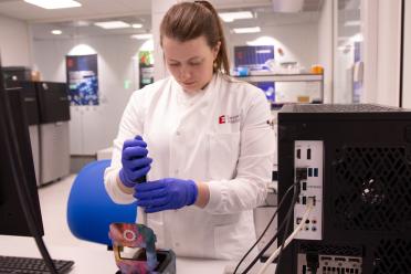 Naomi Irish, wearing a white lab coat and blue safety gloves using the Oxford Nanopore promethion 2 solo sequencer