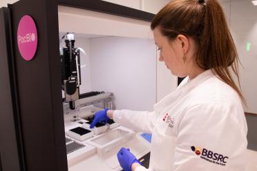 Naomi Irish, wearing a white lab coat and blue safety gloves next to the PacBio Revio sequencer