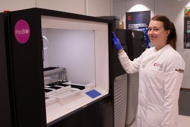 Naomi Irish, wearing a white lab coat and blue safety gloves next to the PacBio Revio sequencer
