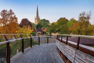 A wooden bridge leading into the distance with the spire of Norwich cathedral in the background